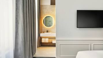 Ett modernt hotellrum med en stor säng, en platt-TV som är monterad på väggen, en rund spegel med en hexagonformad ram och ett badrum med en vit handfat och handduksställ.