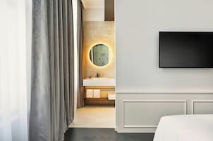 Una camera d'albergo moderna con un letto grande, una TV a schermo piatto montata a parete, uno specchio rotondo con cornice esagonale e un bagno con lavandino bianco e portasci.