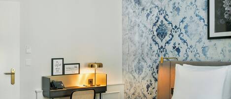 A hotel room with a bed, a desk with a telephone, a chair, and a wall with a patterned wallpaper.