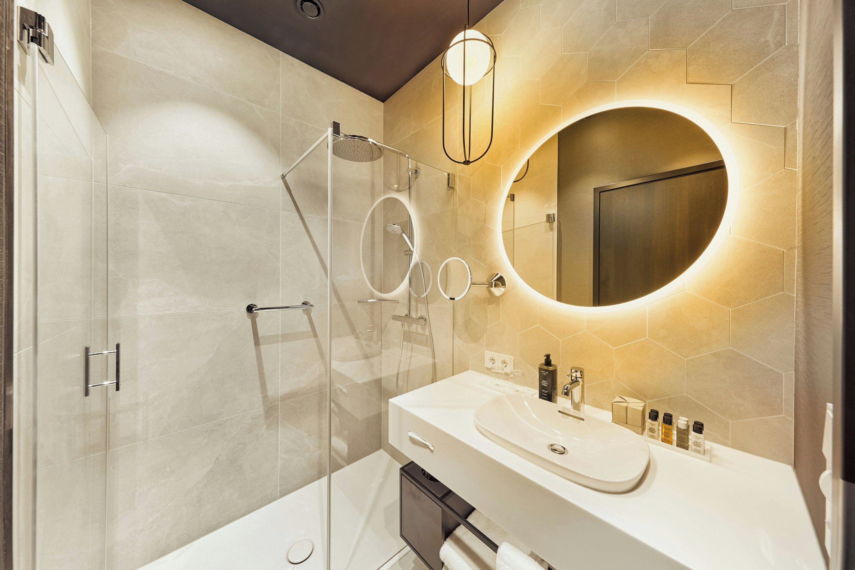 Un baño moderno con cabina de ducha de vidrio, un espejo redondo y un mueble de tocador blanco con artículos de aseo.