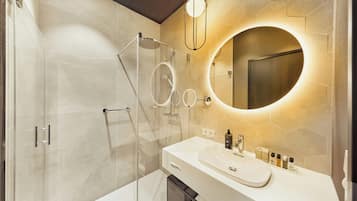 Un baño moderno con cabina de ducha de vidrio, un espejo redondo y un mueble de tocador blanco con artículos de aseo.