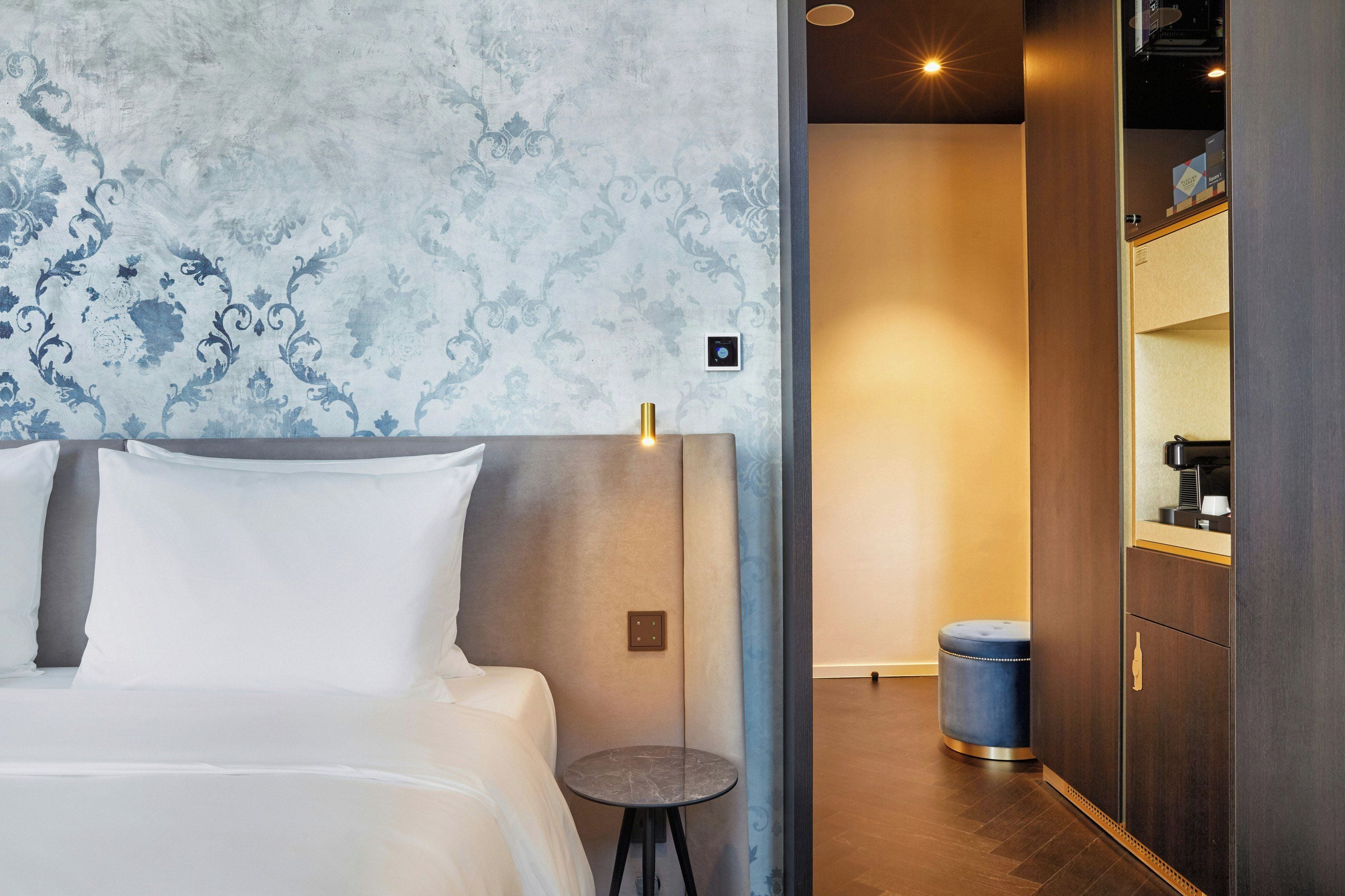 Una habitación de hotel con cama, una mesita redonda pequeña, una mesita de noche, una lámpara y un armario.