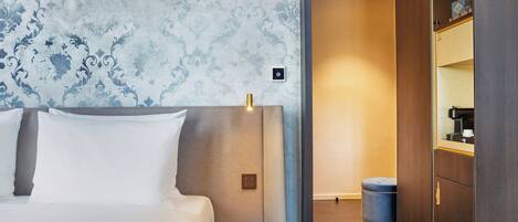 Una habitación de hotel con cama, una mesita redonda pequeña, una mesita de noche, una lámpara y un armario.