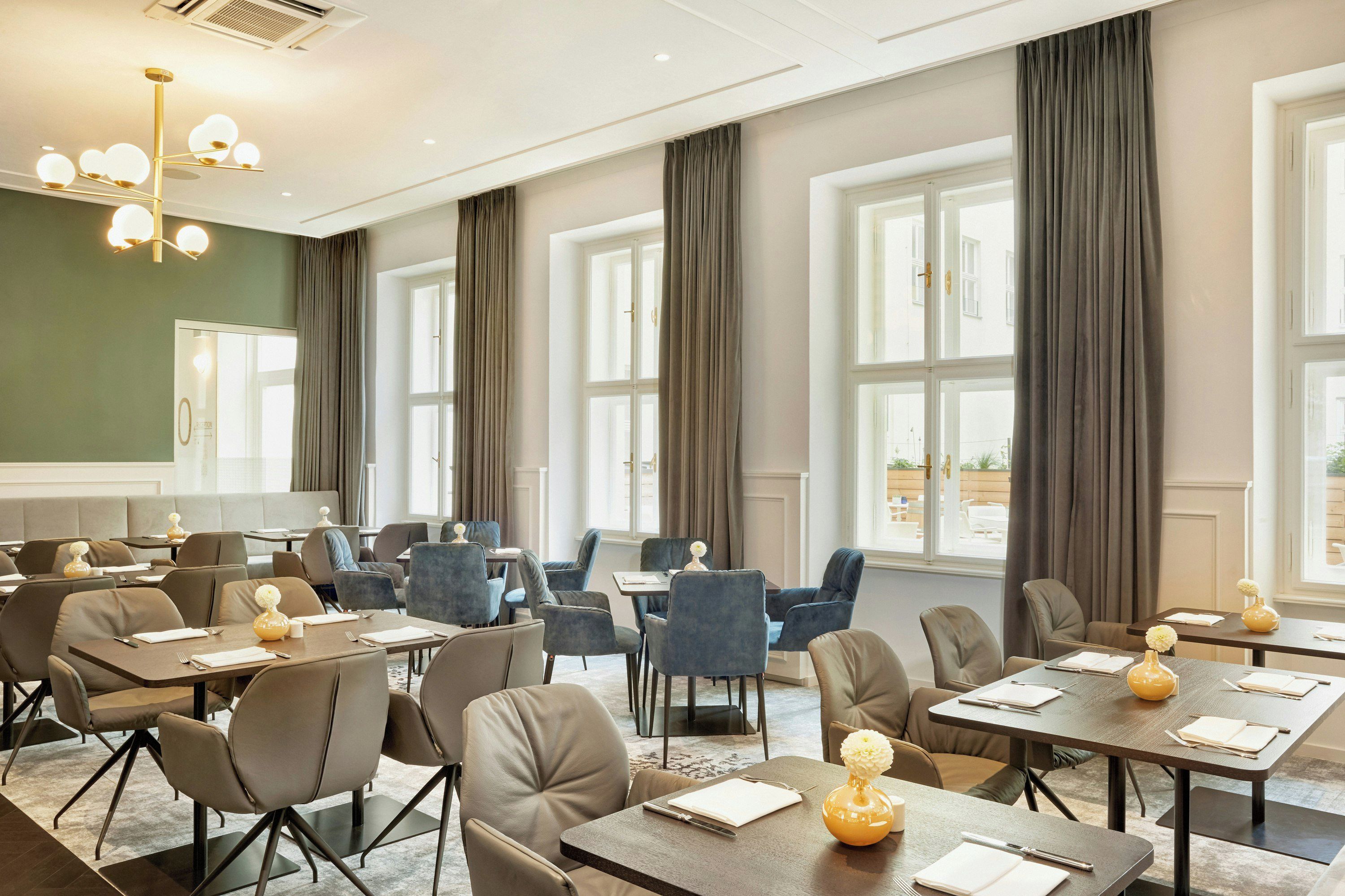 Een hotelconferentieruimte met meerdere tafels en stoelen, grote ramen met gordijnen en een moderne kroonluchter.