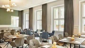Een hotelconferentieruimte met meerdere tafels en stoelen, grote ramen met gordijnen en een moderne kroonluchter.