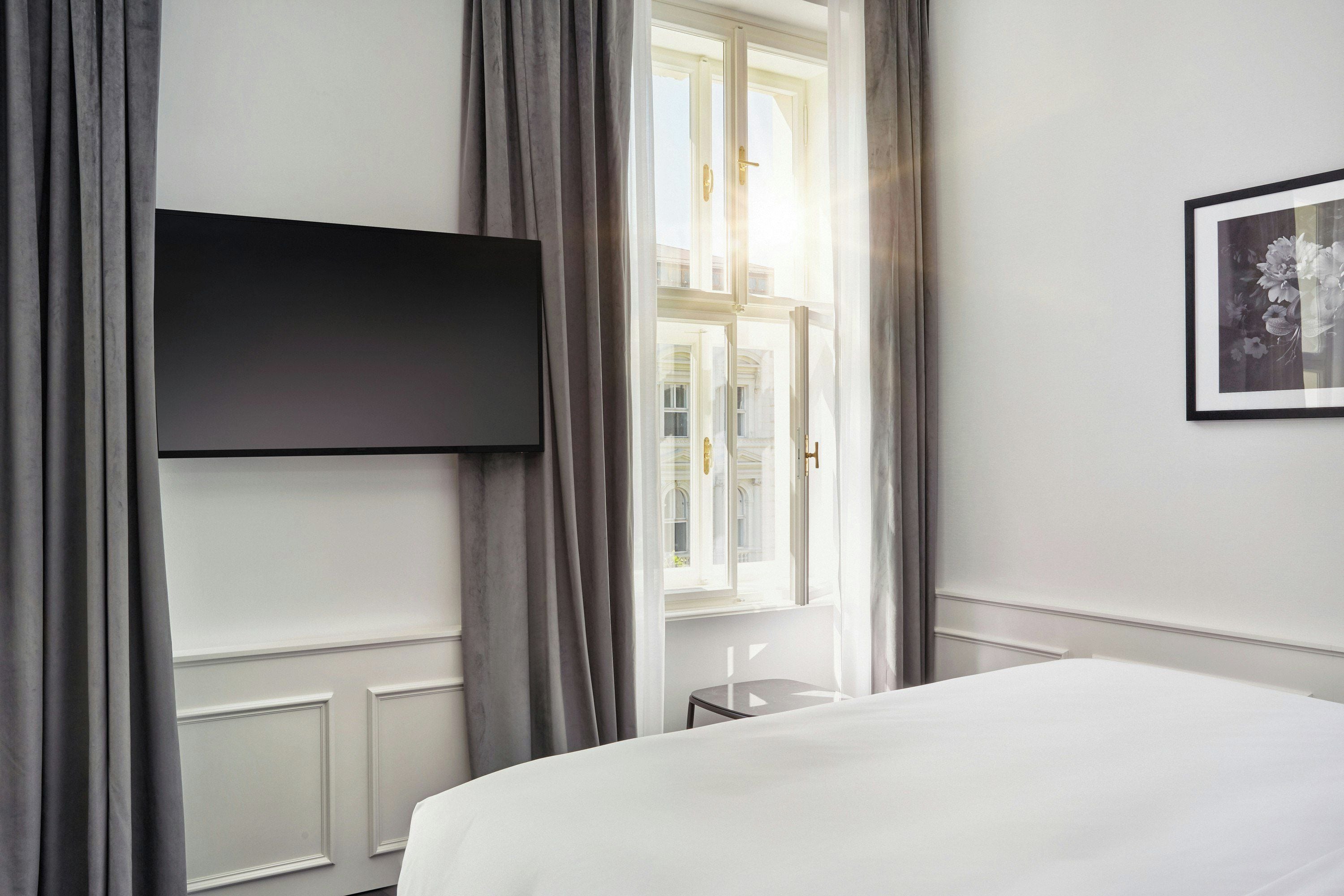 A hotel room with a bed, a flat-screen TV mounted on the wall, a window with curtains, and a framed picture on the wall.