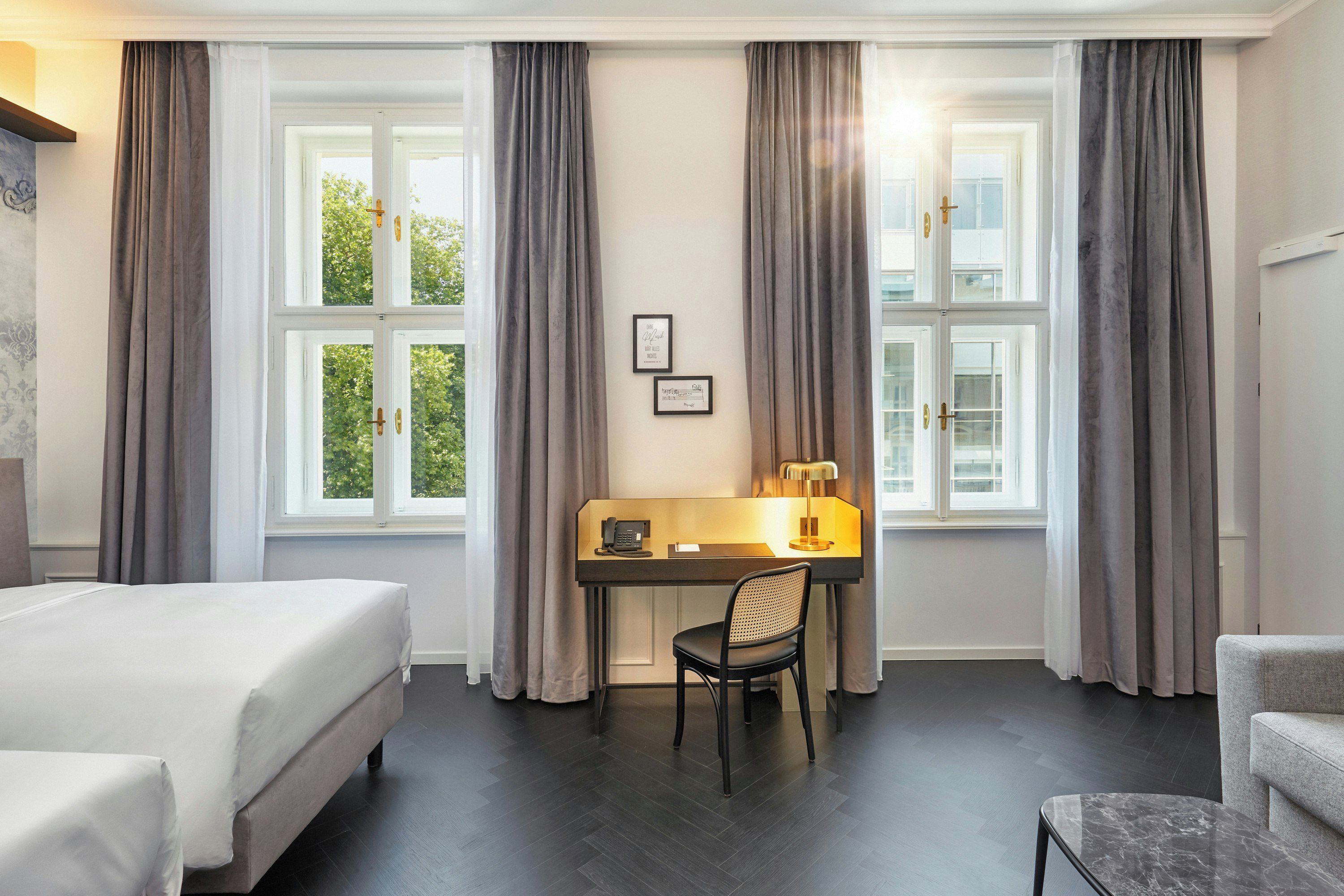 A hotel room with a bed, desk, chair, and large windows with curtains.