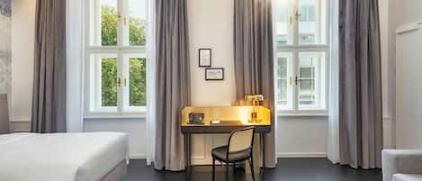 Habitación de hotel con cama, escritorio, silla y ventanas grandes con cortinas.
