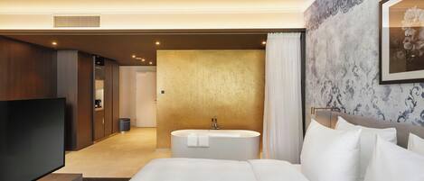 Habitación de hotel con una cama grande, un televisor de pantalla plana, una bañera redonda y un baño con espejo e inodoro.