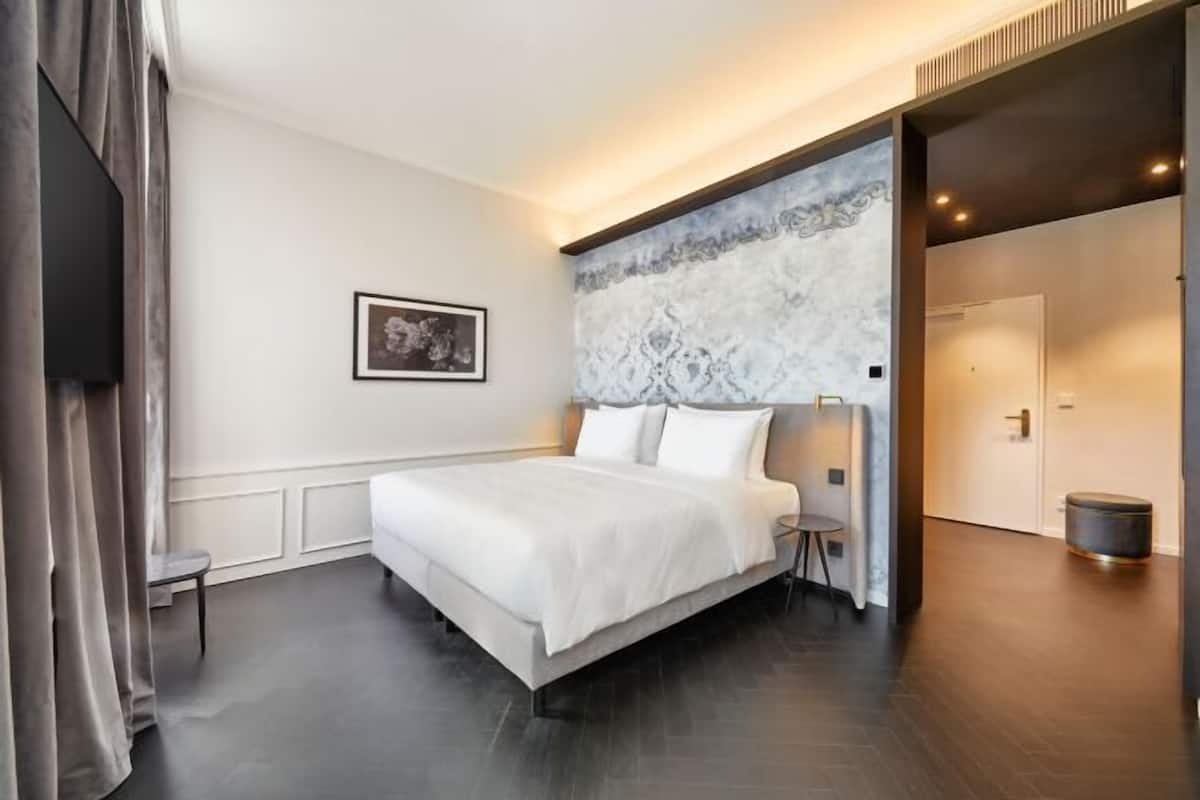 Una habitación de hotel moderna con una cama grande, una mesita auxiliar pequeña, un cuadro enmarcado en la pared y una pared decorativa de mármol.