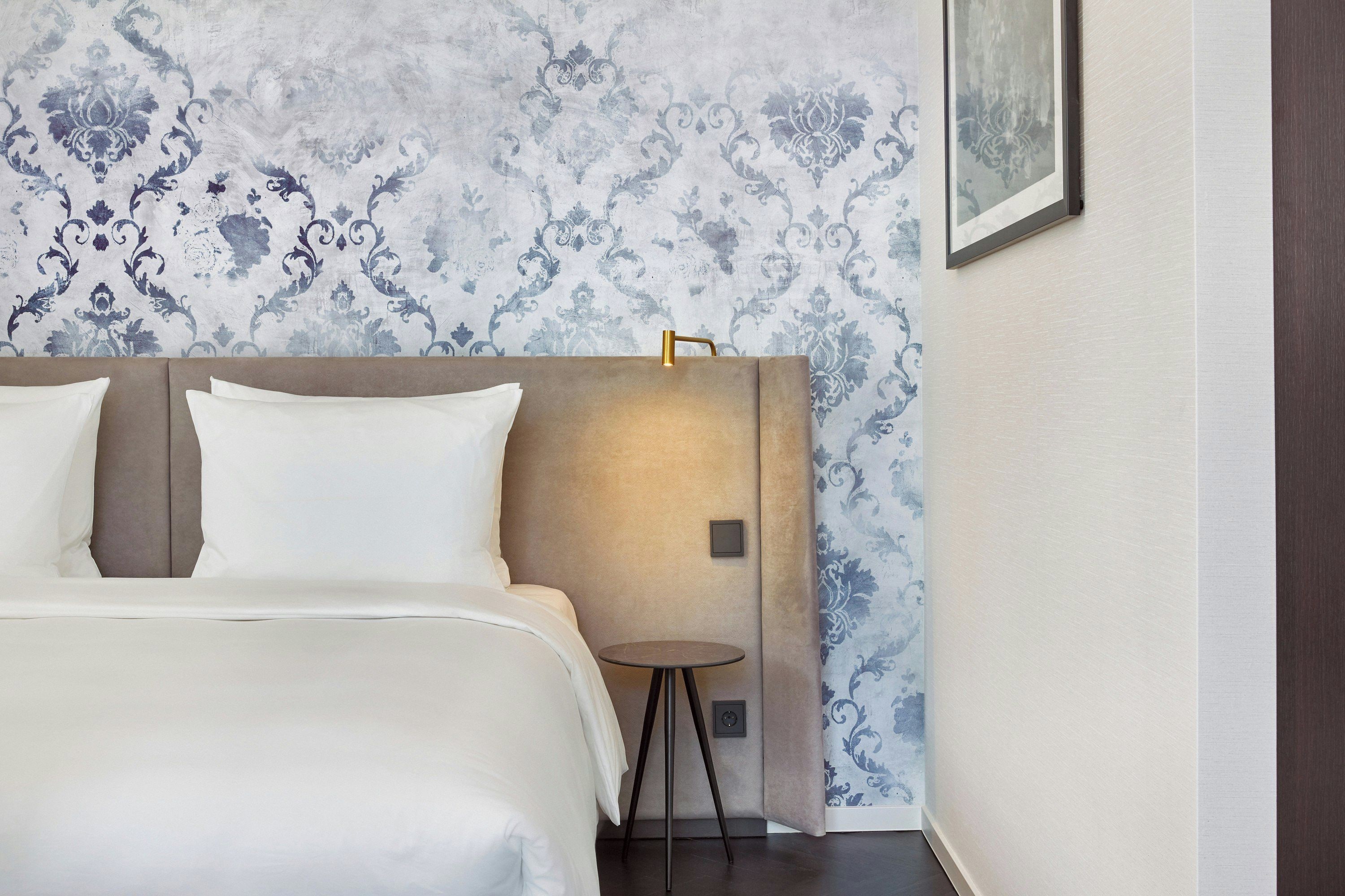 Une chambre d’hôtel avec un lit, une table de chevet, une lampe fixée au mur et un tableau encadré.