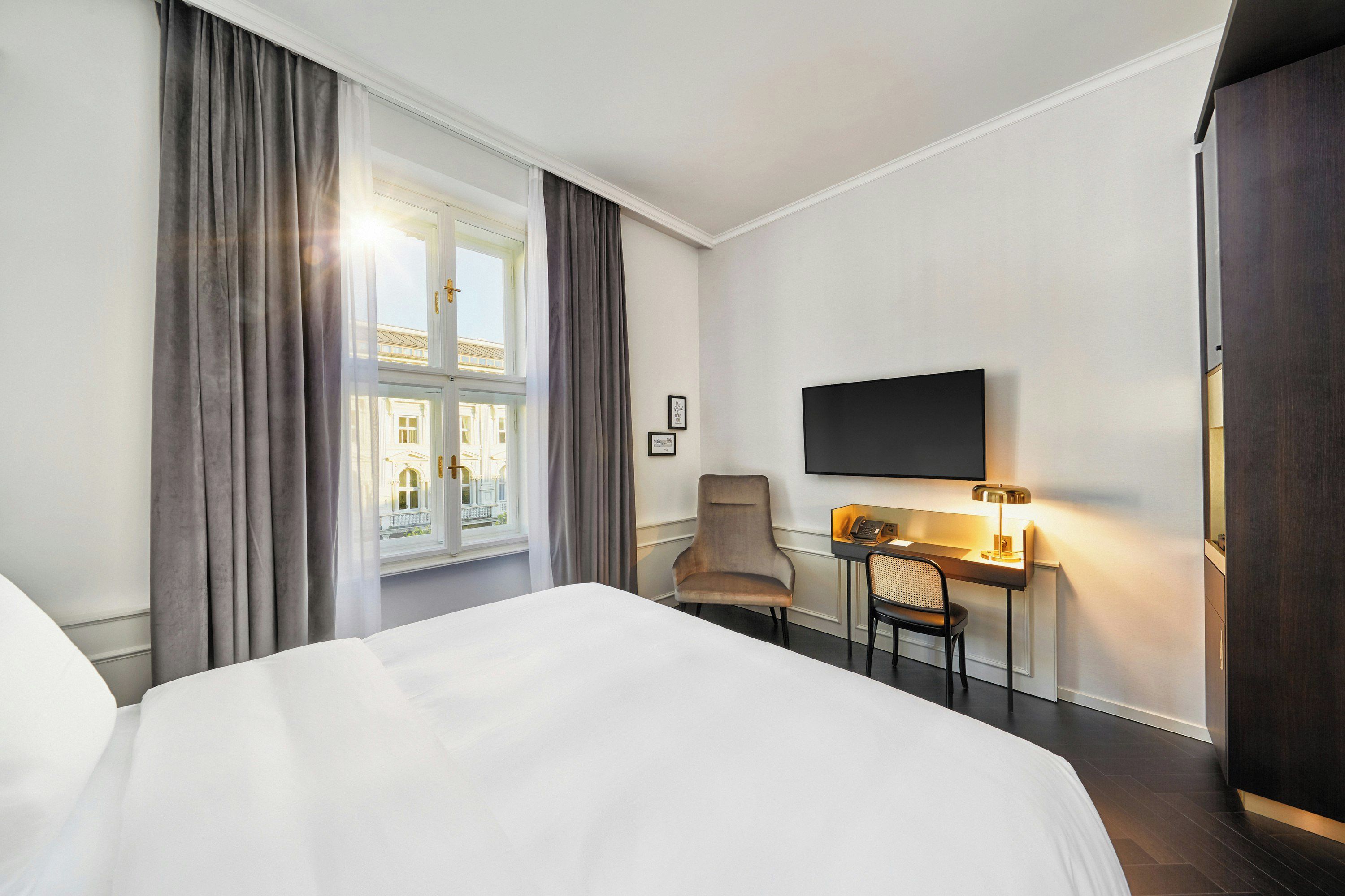 A hotel room with a bed, a desk with a chair, a television, and a window with curtains.