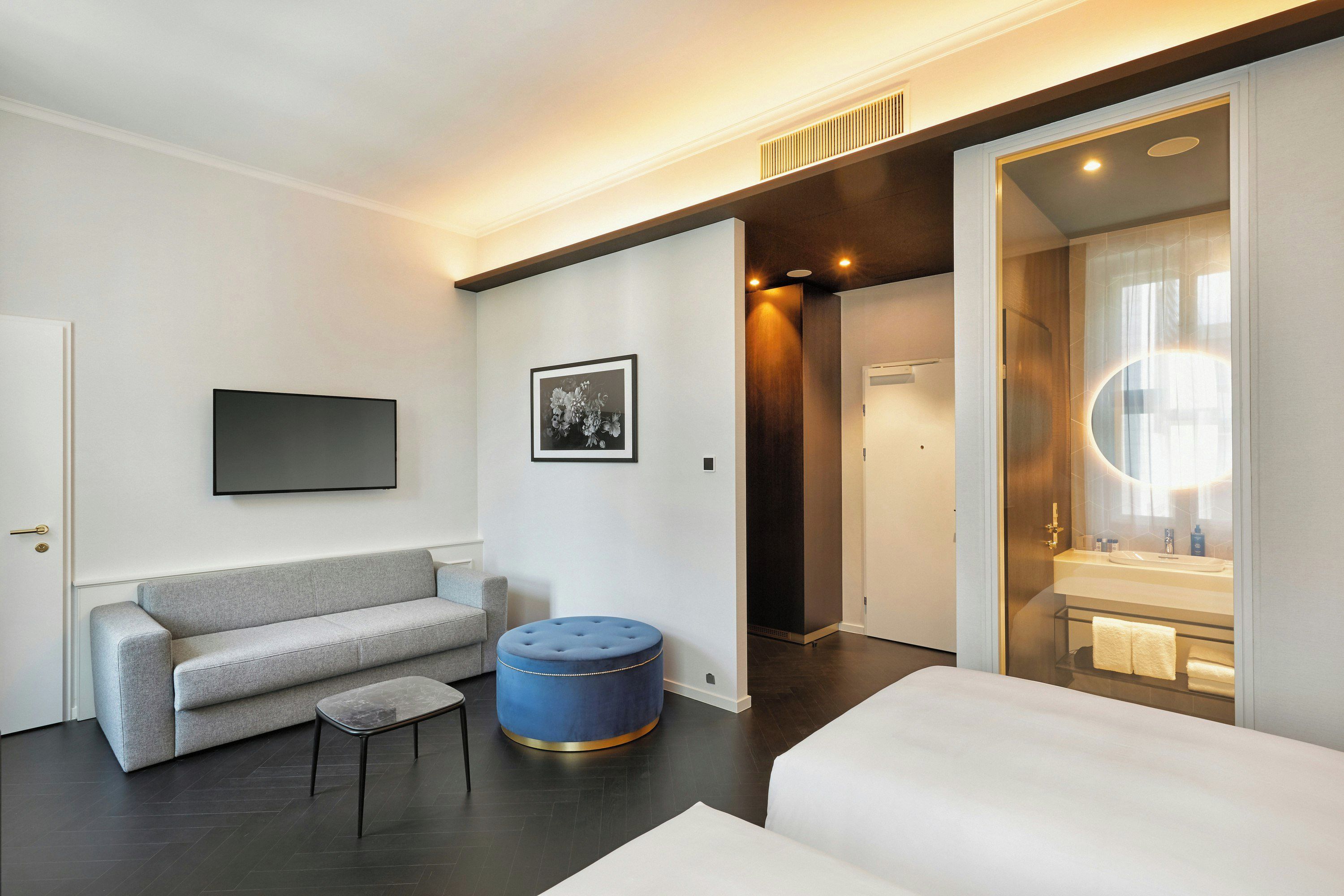 Une chambre d’hôtel moderne avec un canapé gris, un petit pouf rond, un téléviseur à écran plat fixé au mur et une salle de bain visible à travers une porte en verre.