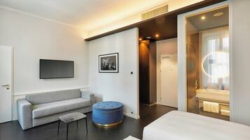 Habitació d'hotel moderna amb un sofà gris, un otomà petit i rodó, una televisió de pantalla plana penjada a la paret i un bany visible a través d'una porta de vidre.