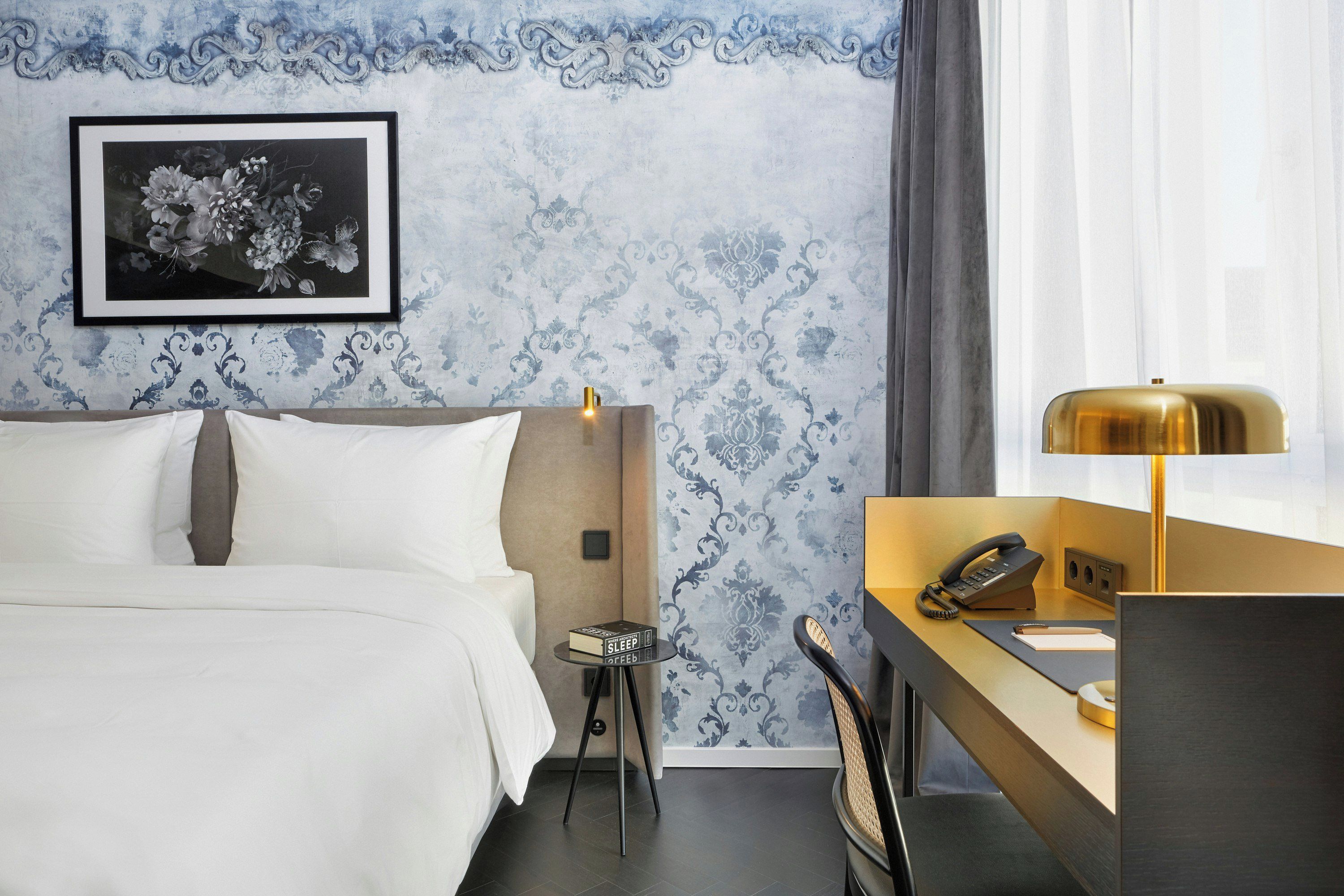 A hotel room with a bed, a desk with a telephone, a small table with a book, and a framed picture on the wall.