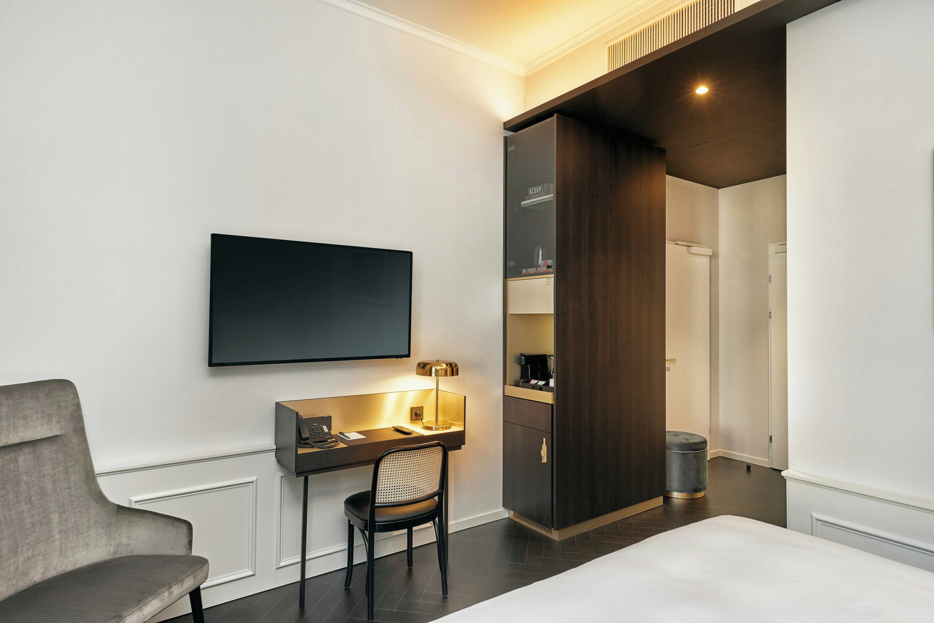 A modern hotel room with a flat-screen TV mounted on the wall, a desk with a lamp, a chair, and a bed.