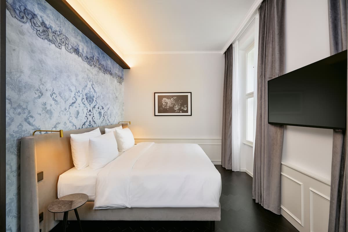Habitación de hotel con una cama grande, una mesita auxiliar pequeña, un televisor montado en la pared y un cuadro enmarcado.