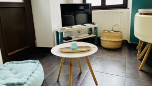 Una habitación con una televisión sobre un mueble bajo, una pequeña mesa redonda con una cesta, un sillón de saco azul claro y una cesta de mimbre en el suelo.