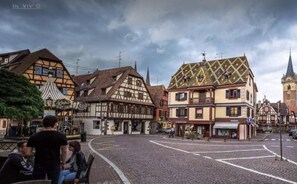 Une place pavée, bordée de bâtiments traditionnels à colombages, avec un beffroi et une fontaine.