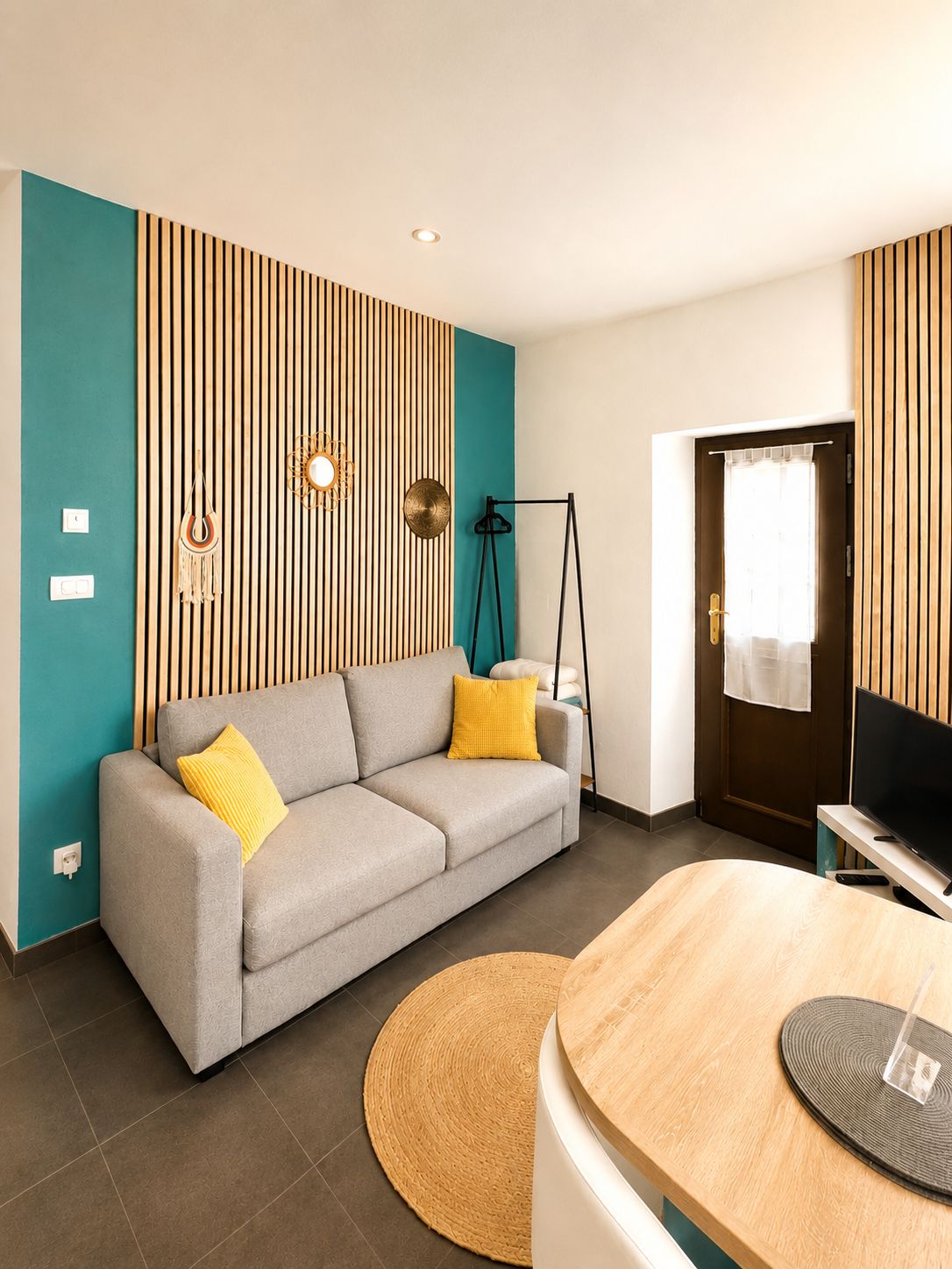 A modern living room with a grey sofa, yellow cushions, a wooden accent wall, a television, and a round wooden table.