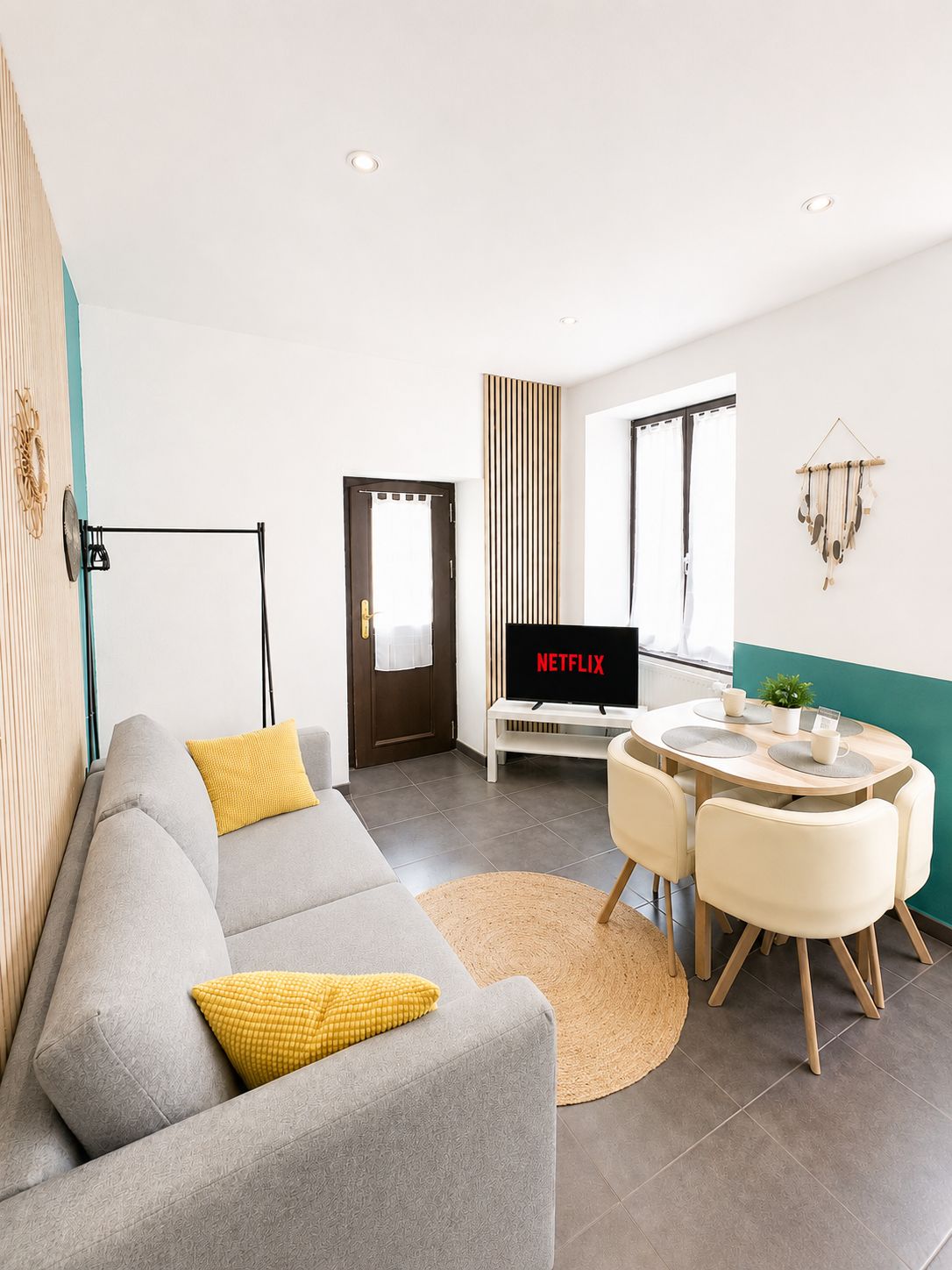 A modern living room with a grey sofa, yellow cushions, a round dining table with chairs, a TV displaying 'NETFLIX', and a wall-mounted decor piece.