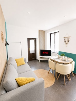 Una sala moderna con un sofá gris, cojines amarillos, una mesa de comedor redonda con sillas, un televisor mostrando 'NETFLIX' y un adorno colgado en la pared.