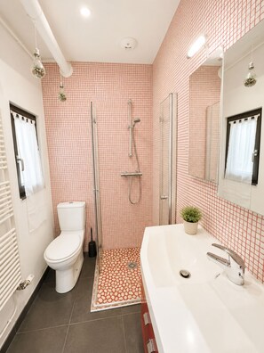 Une salle de bain avec des toilettes blanches, une douche avec une porte en verre, un lavabo avec un miroir et un carrelage décoratif.