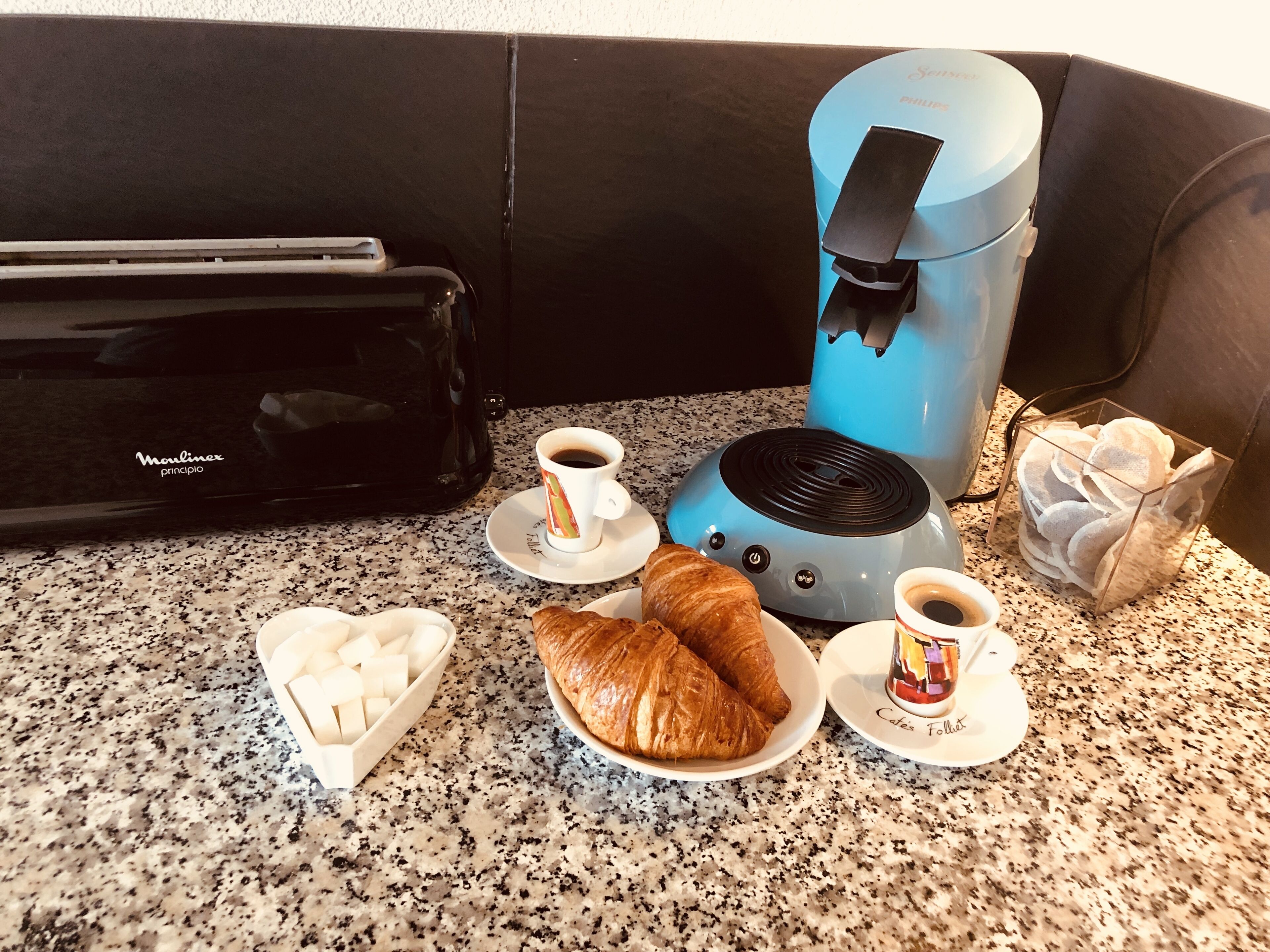 Ein Frühstücksaufenthalt mit einer Kaffeemaschine, Croissants und einer herzförmigen Schale Zuckerwürfel auf einer Granitabdeckung.