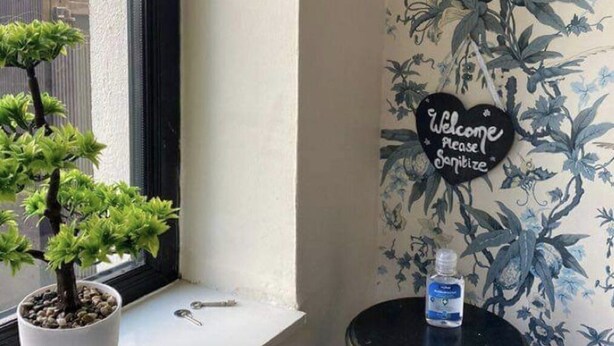 Un bonsai in vaso su un davanzale, un tavolino con una bottiglia, e una parete con carta da parati a motivi floreali e un cartello con su scritto 'Benvenuti, si prega di sanificare'.