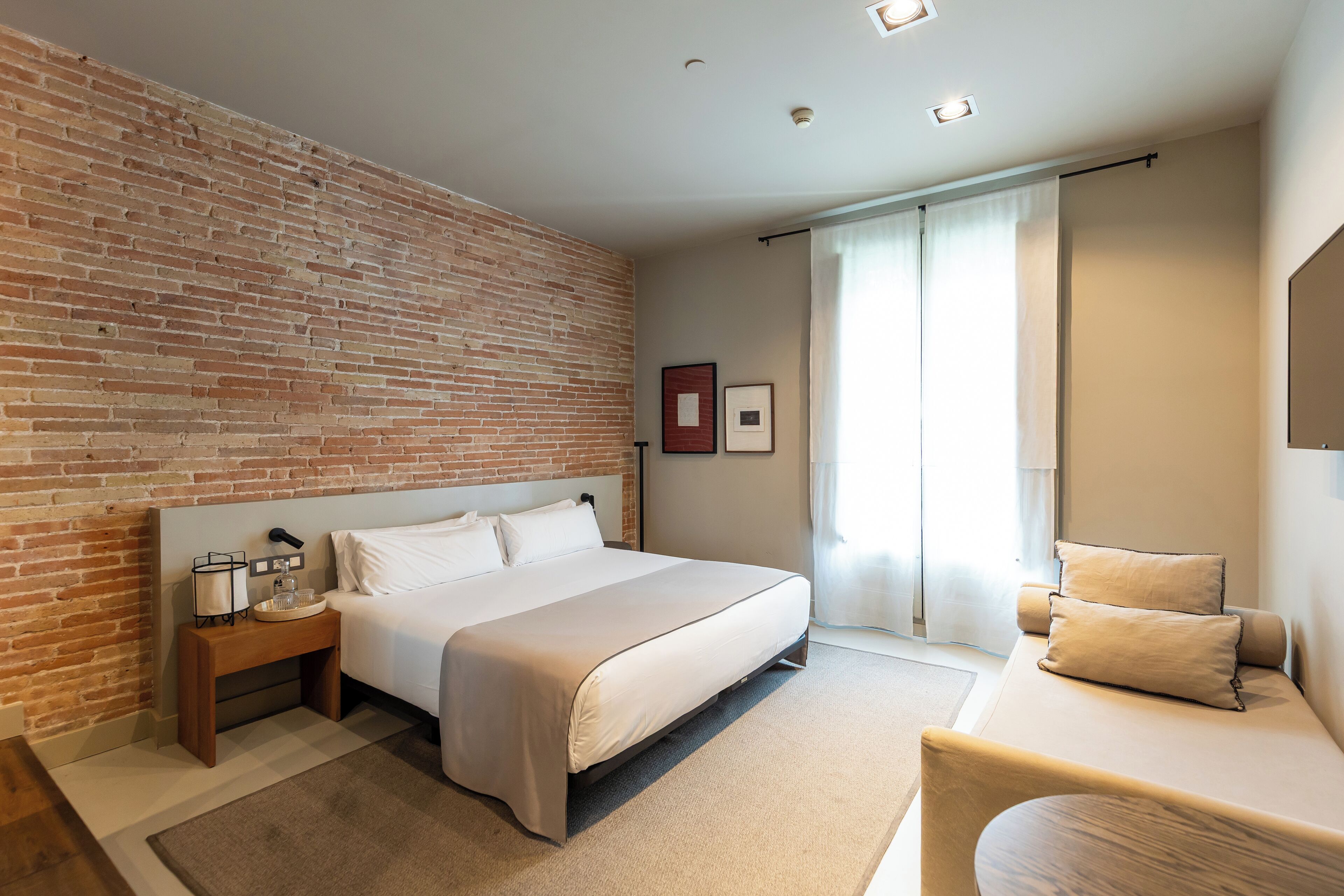 Una habitación de hotel moderna con una pared de ladrillo decorativa, una cama grande, una mesita de noche con una lámpara y vistas al exterior a través de una ventana.