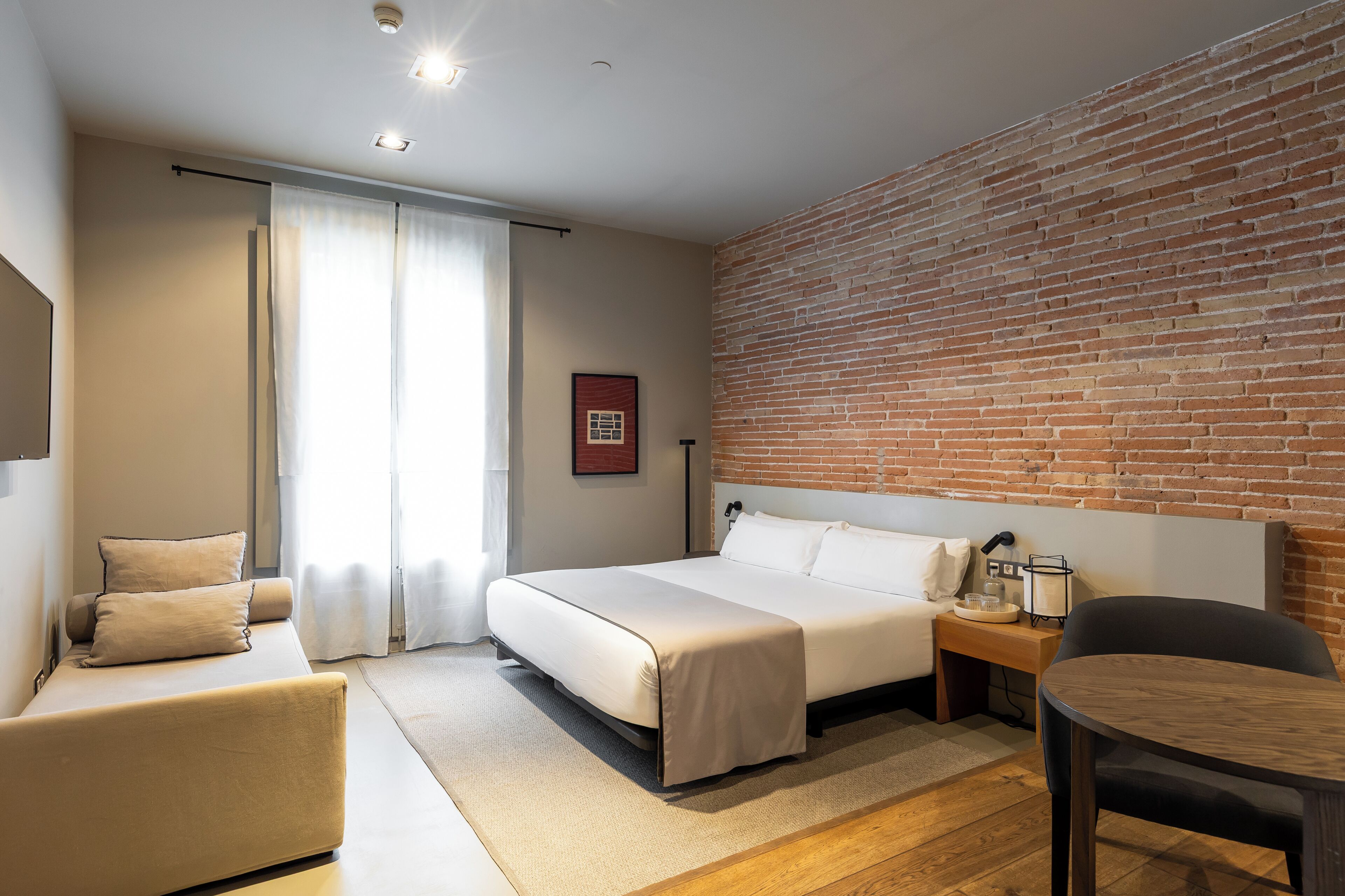Una habitación de hotel moderna con cama, un pequeño sofá, una mesa redonda y una pared de ladrillo.