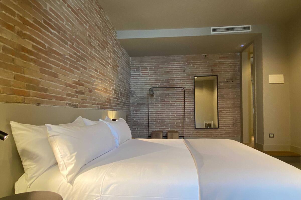 Habitación de hotel con pared de ladrillo, cama grande con ropa blanca, mesita de noche, espejo y puerta que da a otra habitación.