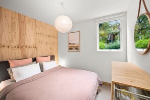 A bedroom with a wooden headboard, a bed with pink bedding, a bedside table, a hanging light fixture, a framed picture, and a window with a view of greenery. - Retro Beach Vibes (Lorne)