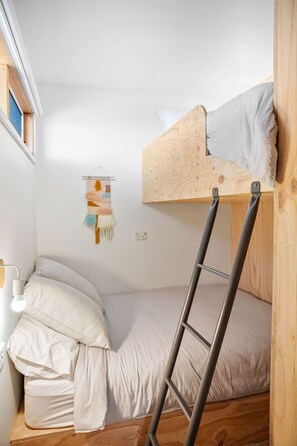 A small, compact room with a bunk bed, a ladder, a wall-mounted shelf with a decorative item, and a bedside table with a lamp.