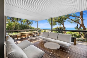 An outdoor deck with a sofa, round table, and benches, overlooking a coastal landscape with trees and the sea. - Retro Beach Vibes (Lorne)
