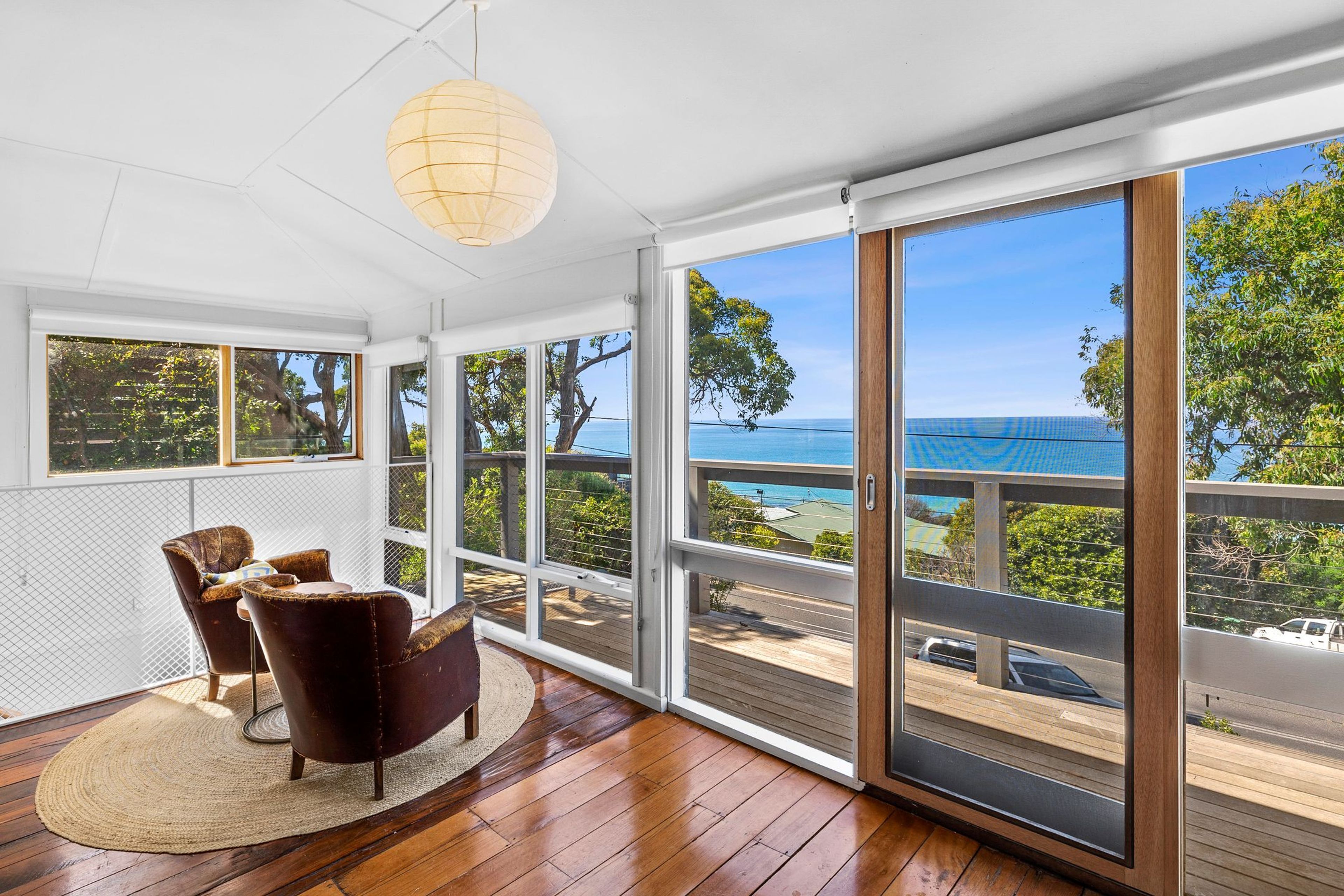 A room with a large sliding glass door leading to a deck with a view of the ocean. There is a round table with chairs and a hanging light fixture.