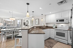 A modern kitchen with white cabinetry, granite countertops, stainless steel appliances, pendant lighting, and a view of the city.
