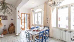 Uma sala de jantar com uma mesa posta para quatro pessoas, um candelabro e vista para a rua através de grandes janelas.