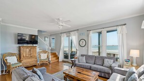 Un salon comprenant un canapé, une table basse, des fauteuils en osier, un téléviseur, une horloge et une vue sur l’océan grâce à de grandes fenêtres.