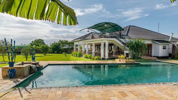 A pool area with a covered patio, a white house with a balcony, and a well-maintained lawn. - Moyoni Airport Lodge (Arusha)