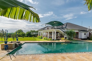 A pool area with a covered patio, a white house with a balcony, and a well-maintained lawn. - Moyoni Airport Lodge (Arusha)