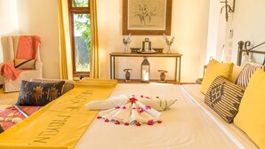 A hotel room with a neatly made bed, a sitting area with a chair and cushions, a small table with decorative items, and a window with curtains. - Moyoni Airport Lodge (Arusha)