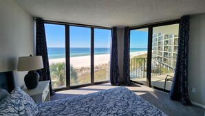 Un dormitorio con un ventanal grande con vistas a la playa, una cama con ropa de cama estampada y una mesita de noche con una lámpara.