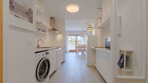 Een smalle keuken met witte wanden, een wasmachine, een eethoek met tafel en stoelen en een uitzicht door een glazen deur naar buiten.