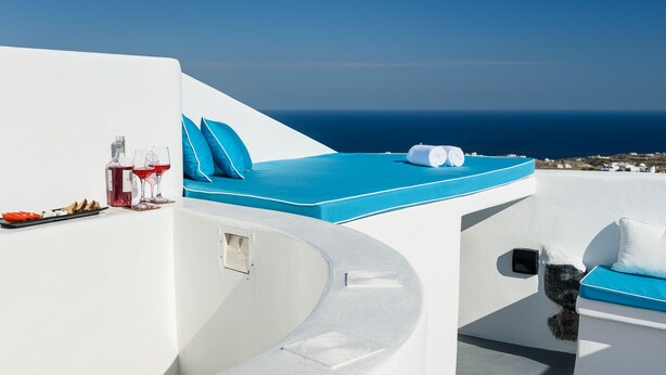 Un área de estar exterior con asientos blancos y azules, vista al mar, una botella de vino tinto y una copa.