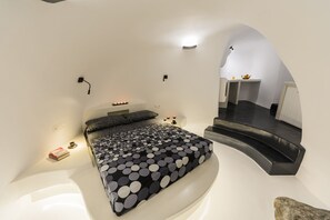 Un dormitorio moderno con una cama curva, una mesita de noche pequeña y una cómoda. Se ve una escalera que sube a un nivel superior.