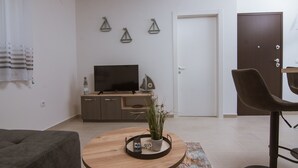 A modern living room with a sofa, a round wooden coffee table with a remote control and a potted plant, a TV on a wooden cabinet, and a door in the background. - Apartments PiiP-ni-More (Vodice)