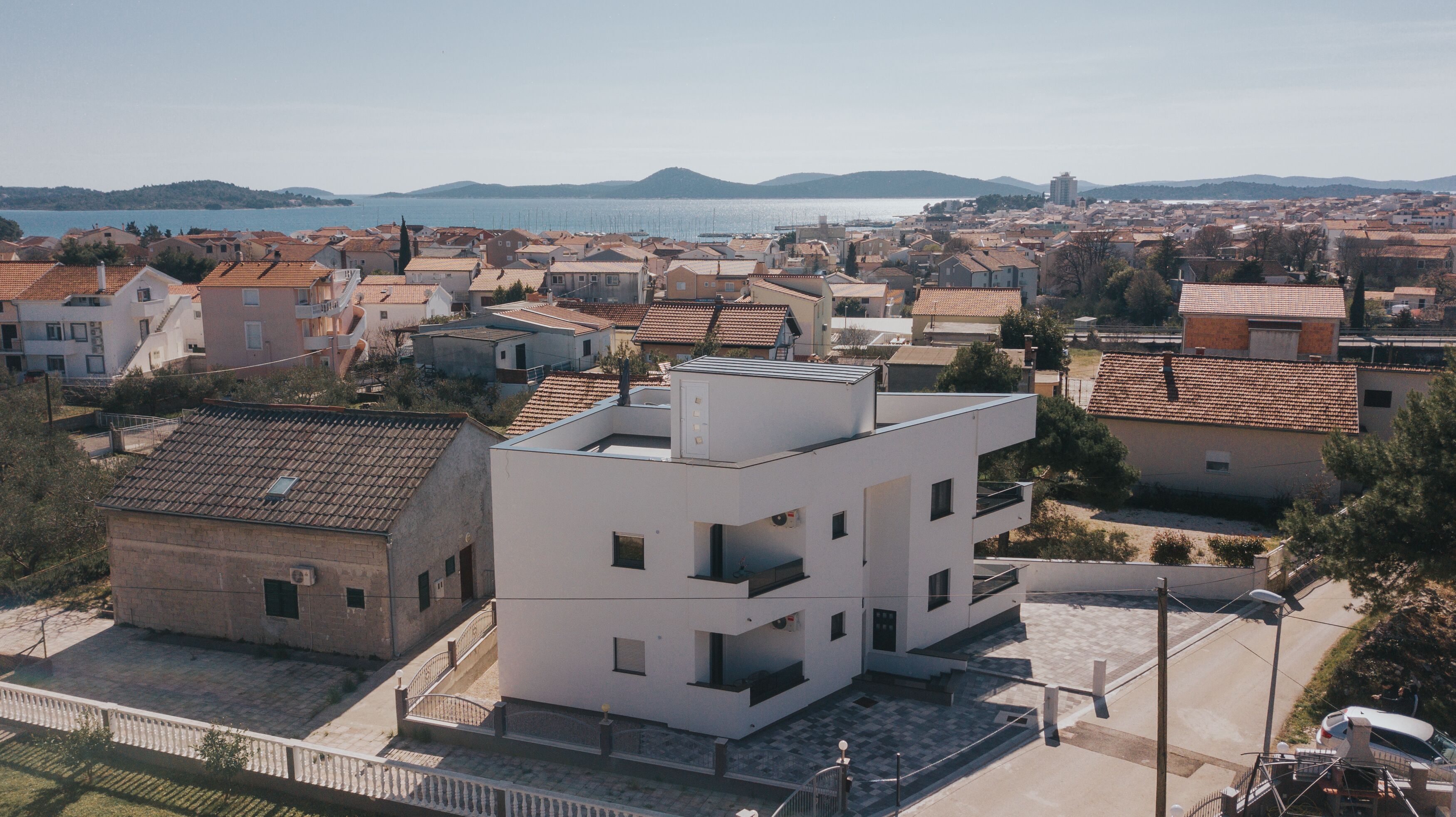 A modern building with a flat roof and a balcony, surrounded by traditional houses with red-tiled roofs.