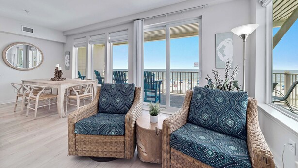A modern living room with wicker furniture, a round table, and a view of the beach through large windows.