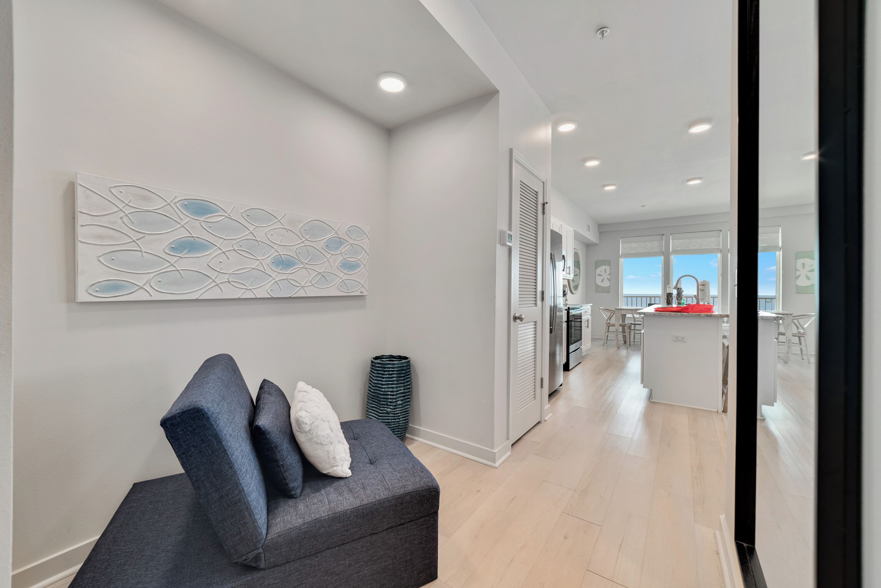 A modern living room with a grey sofa, a decorative wall piece, and a view into the kitchen area.